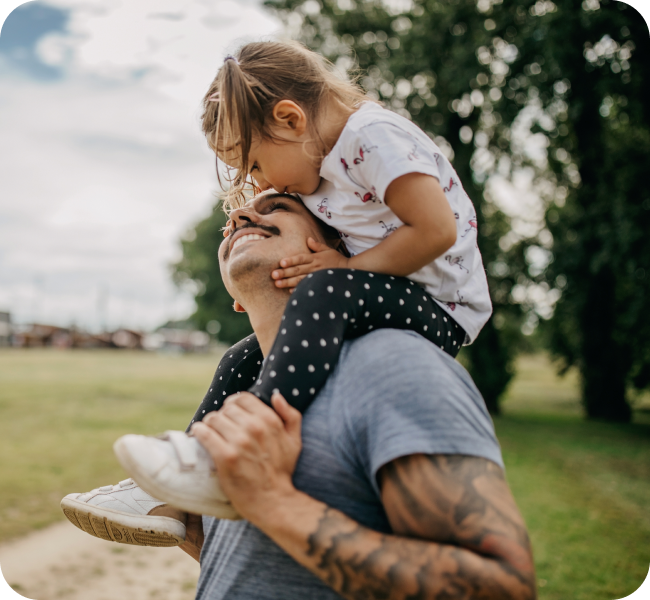 A happy image of a daughter riding on her dad’s shoulders and giving him a kiss on the cheek.