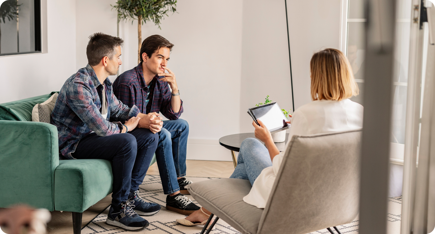  Two men on a couch holding hands meeting with a counselor in a professional setting. 