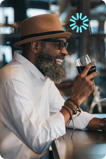 A close up picture of a dapper man in a hat smiling as he takes a sip of wine.